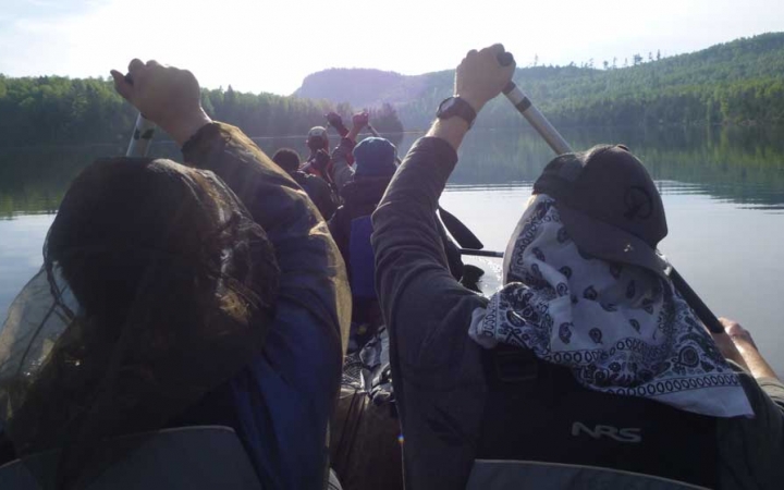 From the back of a canoe, you can see the people in front paddling. The boat is on calm water with trees lining the shore. 